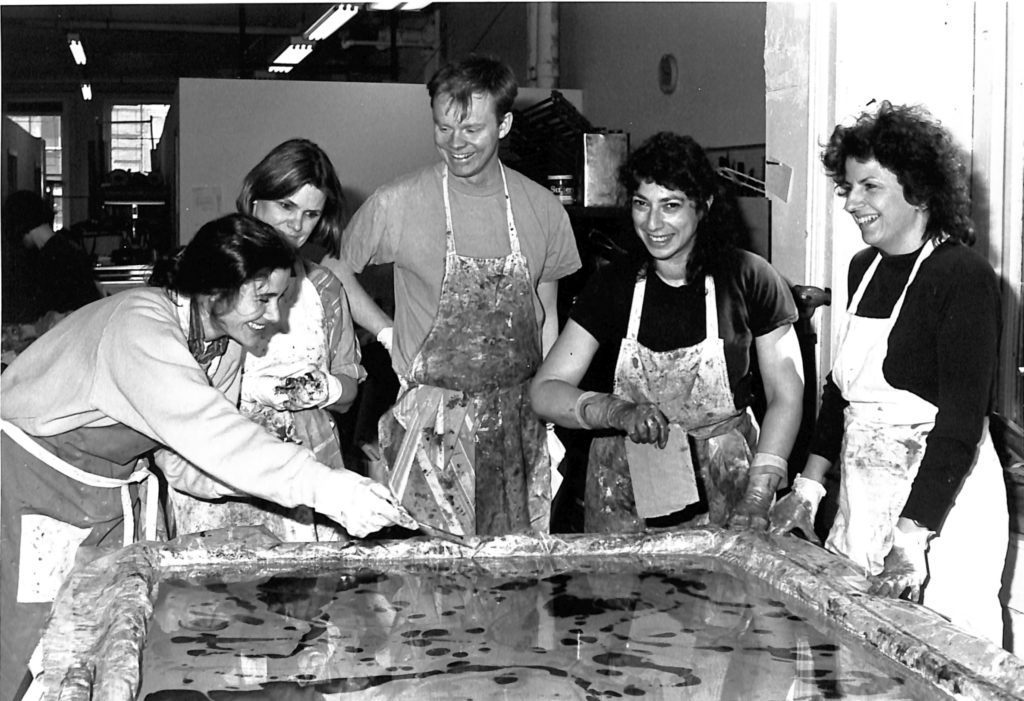 A black and white image of four women and one man in stained aprons working over a wide tray of water and ink to marble paper. They are all smiling.