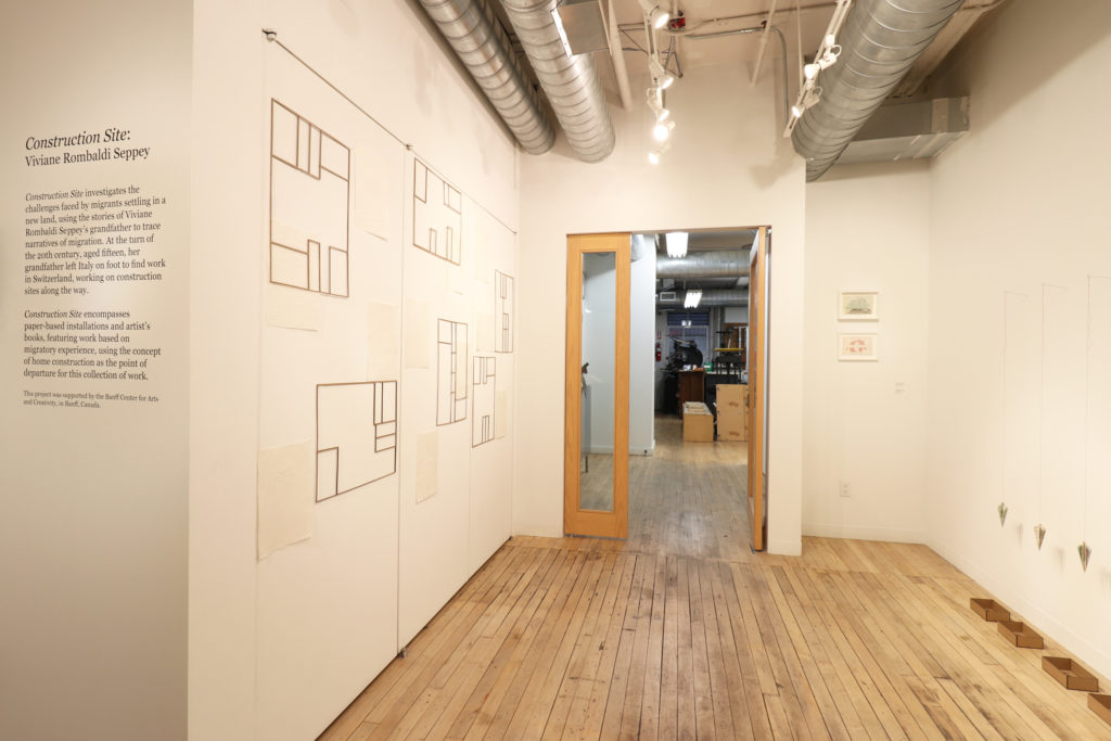 Installation view of Construction Site by Viviane Rombaldi Which has hanging artworks on both sides of the image, a vinyl description on the far left, and a door into the bindery straight ahead.