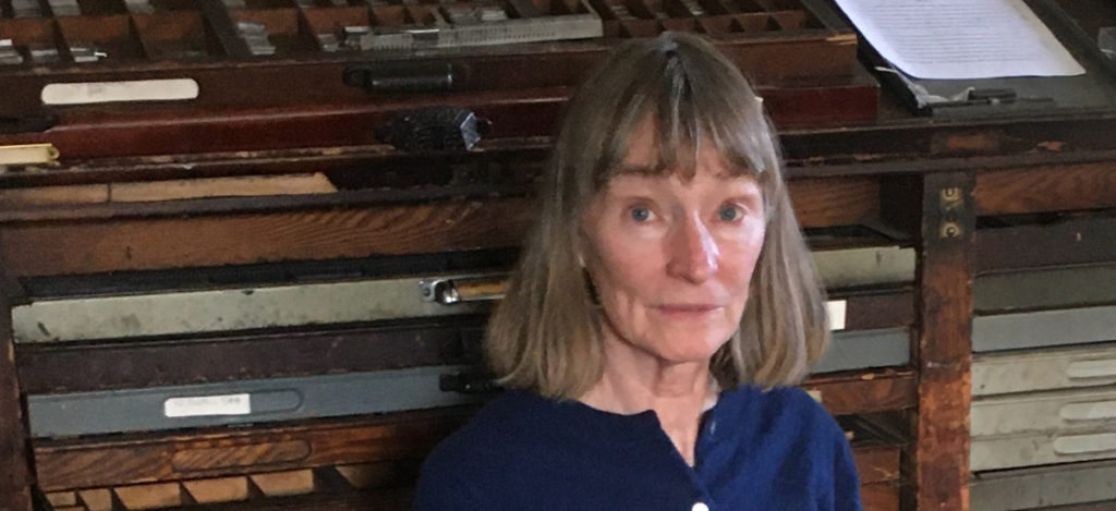 Woman with straight gray hair stands in front of type cases